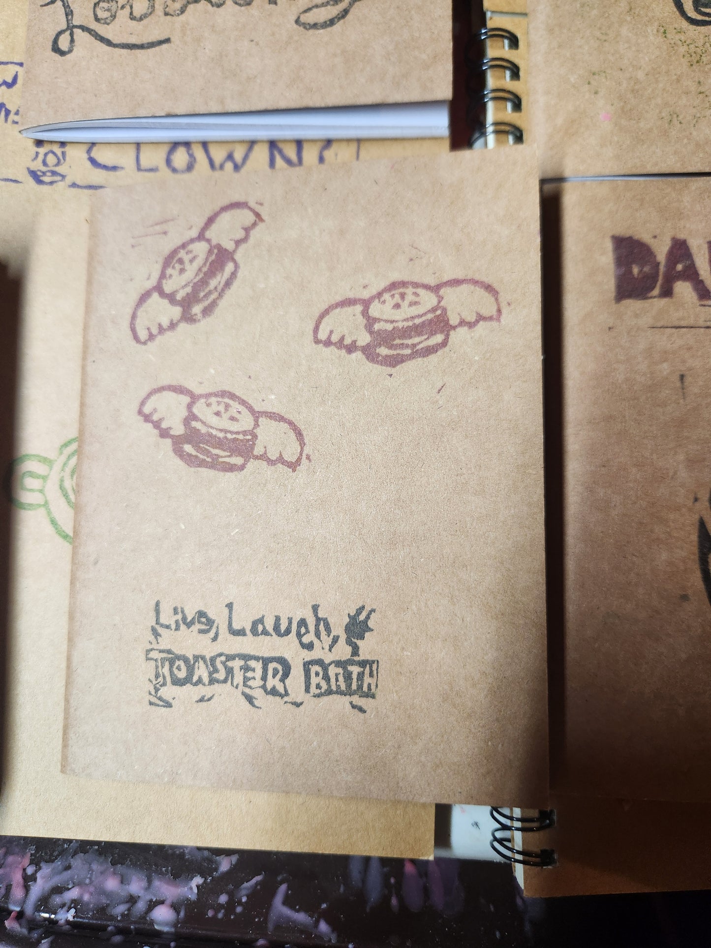 Close up of the cover of a stamped craft paper note book.
Features three flying hamburgers, with live laugh, toaster bath, stamped at the bottom